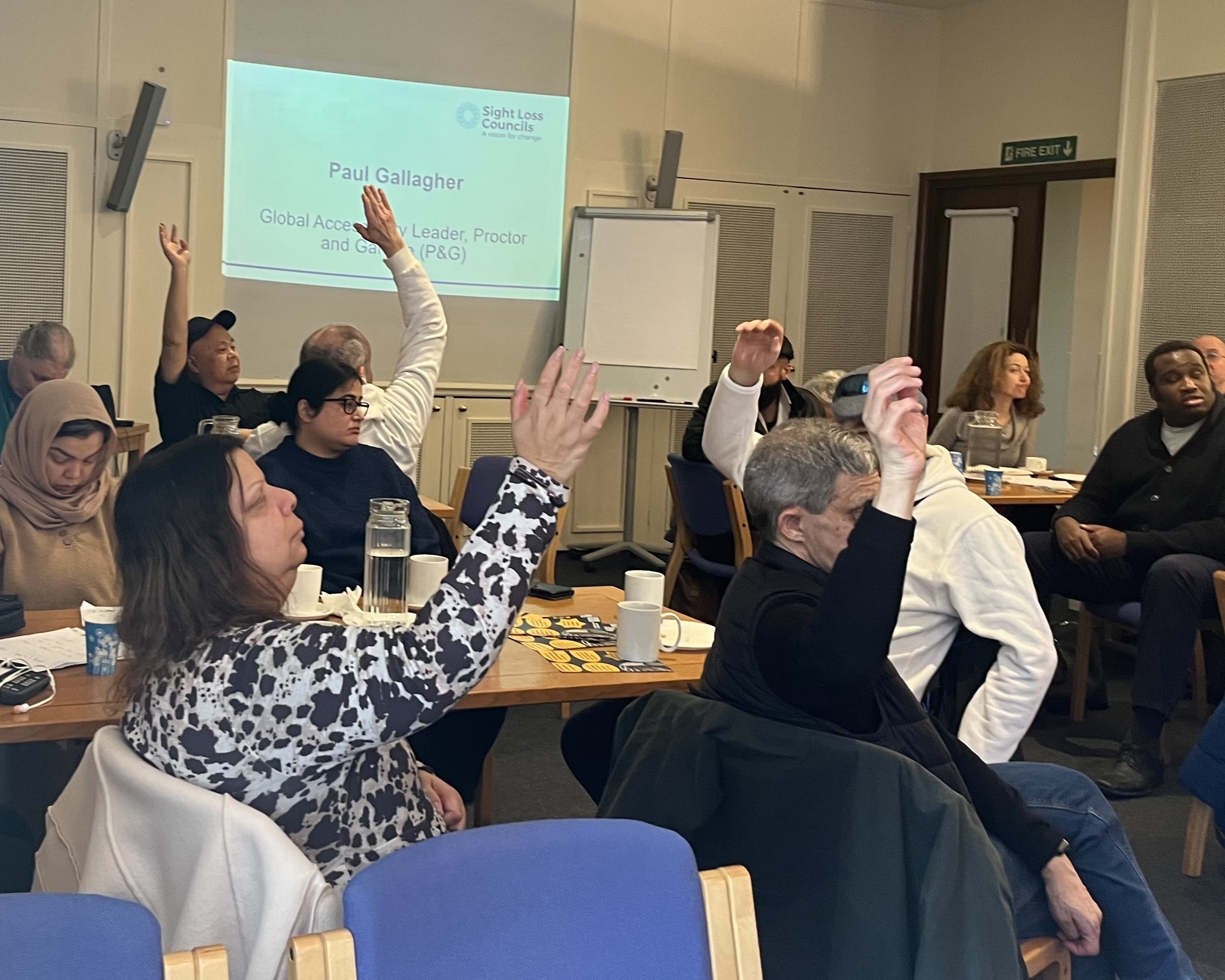 Image looks across the room of delegates, most of whom have their hand raised, during the retail session. In the background an overhead projector screen reads Paul Gallagher, Global Accessibility Leader, Procter and Gamble.