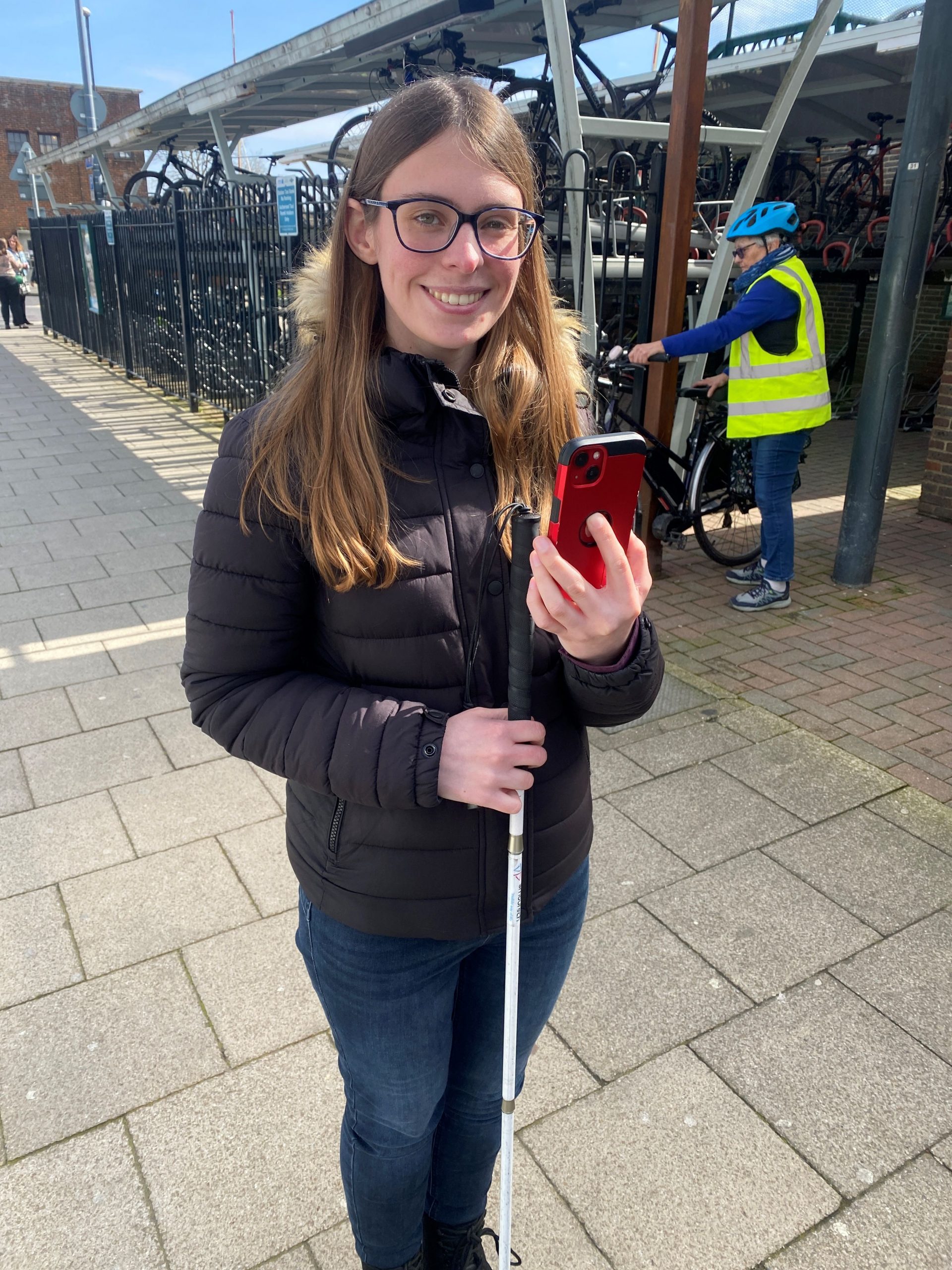 Lauren, SLC coordinator for South East England, pictured at Chichester station. She is holding her cane in one hand, and smart phone in the other. She is smiling at the camera.