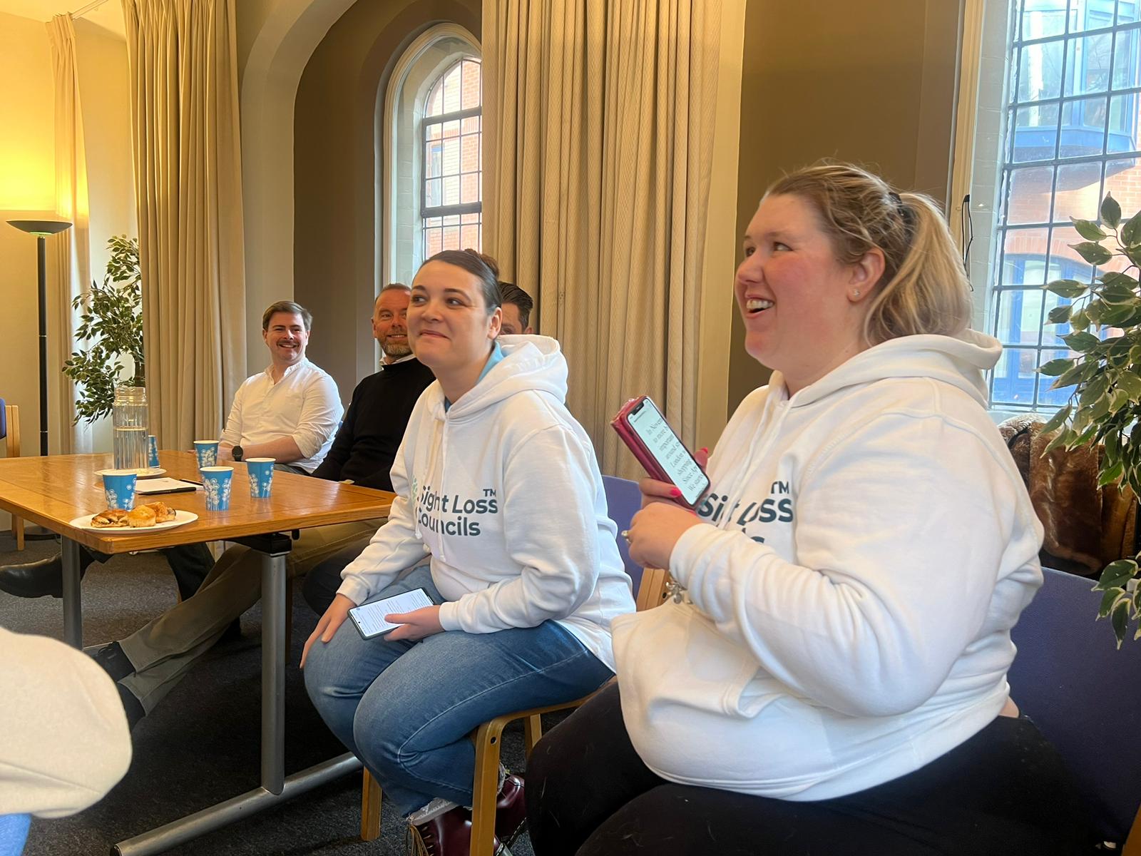 Leanna Best, SE London SLC member, and Nikki Hughes, SW London SLC member, sat talking and laughing with someone off camera. In the background, guest speakers can be seen sitting at a table.