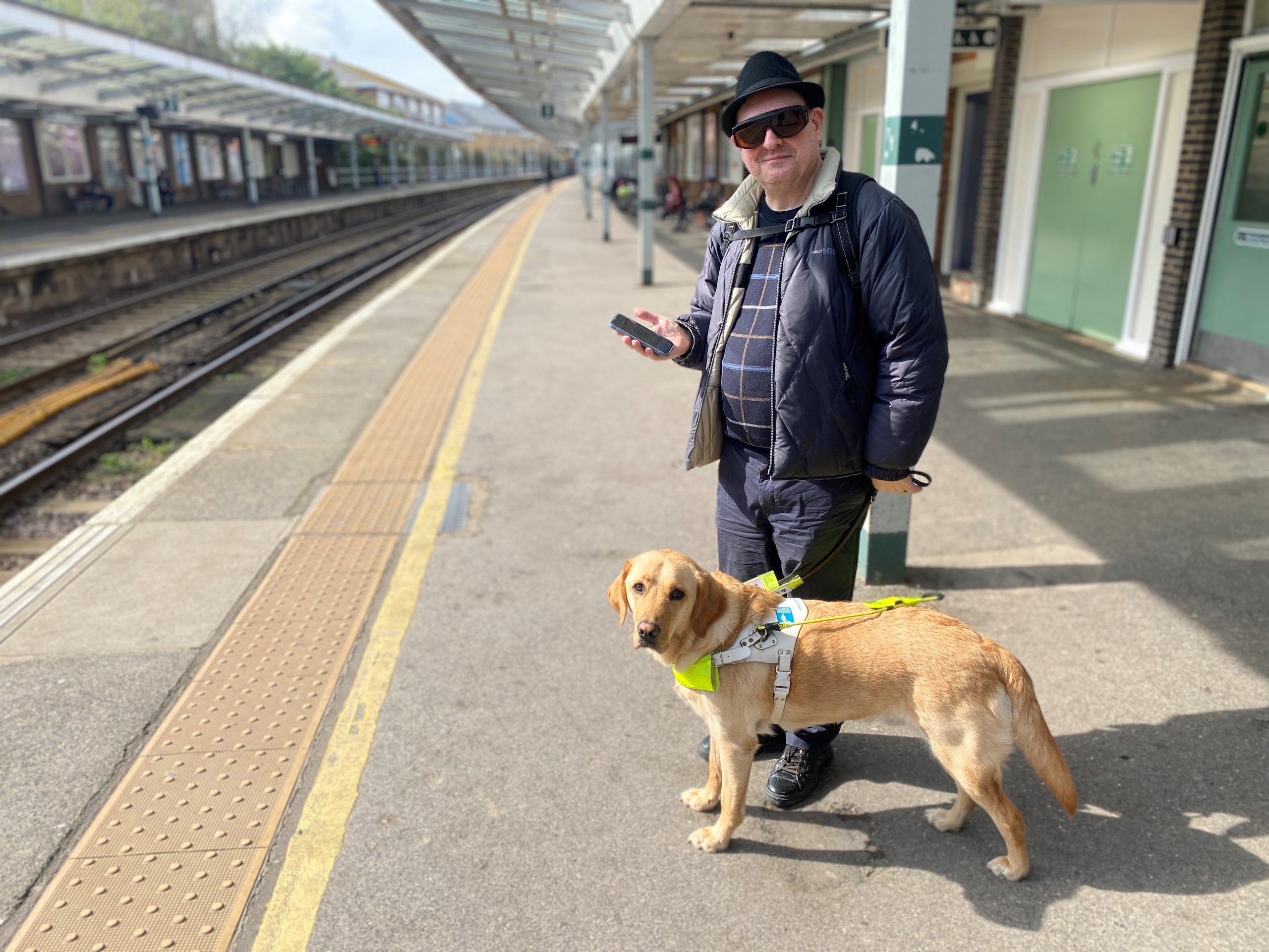 Paul Goddard and his Guide Dog Stevie, picture on a platform at Chichester Station. Paul is holding his smart phone in his hand, smiling at the camera.
