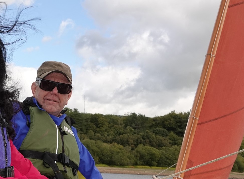 Michael Parkinson - Lancashire SLC member, sitting on a sail boat. To the right of the image, a glimpse of an orange sail can be seen.