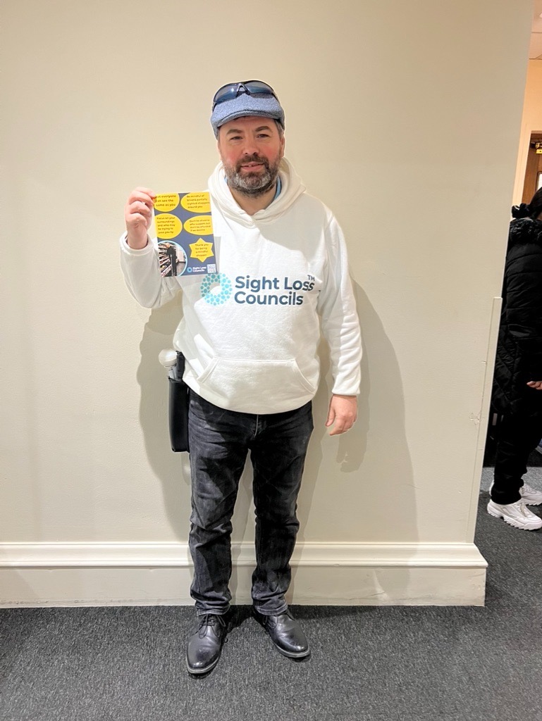SE London SLC member, Steve Reed, pictured holding London SLCs' 'Mindful Shopper' poster which they designed as part of their retail work. He is standing against a cream wall, wearing a white Sight Loss Councils hoodie.