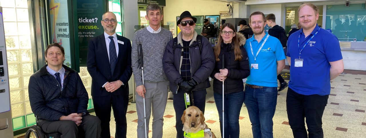 From left to right: Carl Martin, Accessibility Lead at GTR, Dave Smith, Engagement Manager for South East England, Paull Goddard, East Sussex SLC member, Lauren Eade, SLC coordinator, with staff members from GTR. They are stood together in a line in the ticket hall at Chichester Station.