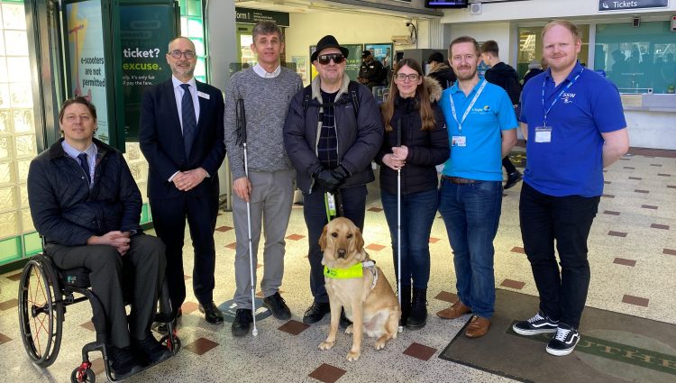 From left to right: Carl Martin, Accessibility Lead at GTR, Dave Smith, Engagement Manager for South East England, Paull Goddard, East Sussex SLC member, Lauren Eade, SLC coordinator, with staff members from GTR. They are stood together in a line in the ticket hall at Chichester Station.