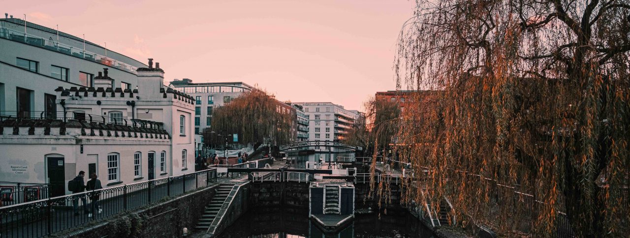 Landscape image of Camden Lock at sunset. A white building is shown to the left of the canal, and branches of a large tree are hanging over the canal on the right. The sky is pink and the canal is full of reflections from its surroundings.