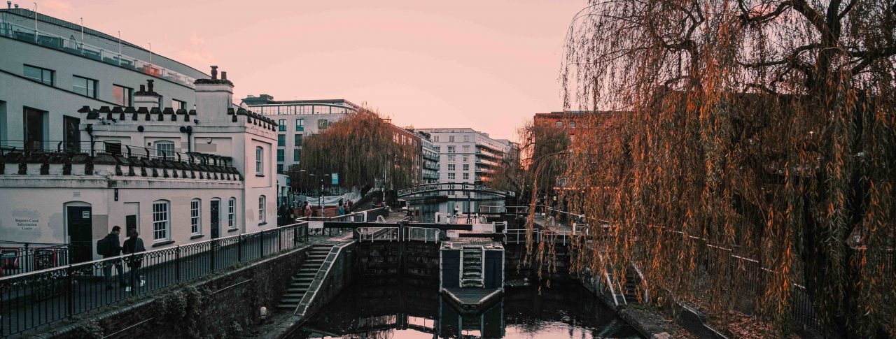 Landscape image of Camden Lock at sunset. A white building is shown to the left of the canal, and branches of a large tree are hanging over the canal on the right. The sky is pink and the canal is full of reflections from its surroundings.