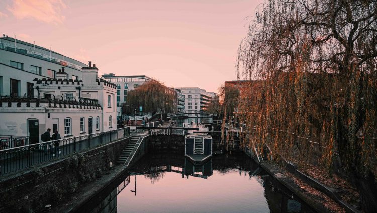 Landscape image of Camden Lock at sunset. A white building is shown to the left of the canal, and branches of a large tree are hanging over the canal on the right. The sky is pink and the canal is full of reflections from its surroundings.