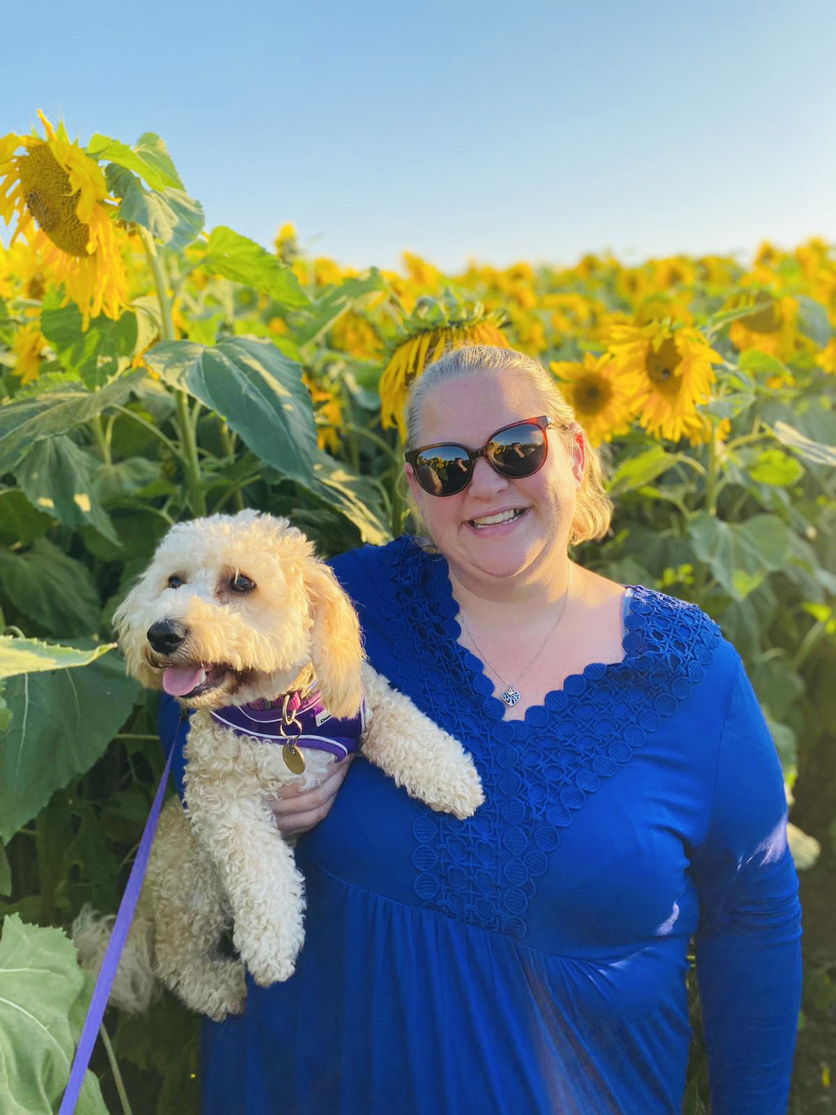 Dannie Gage, Derbyshire SLC member. Dannie is standing amongst a field of towering sunflowers, holding a small fluffy dog, under her arm. She is smiling at the camera.