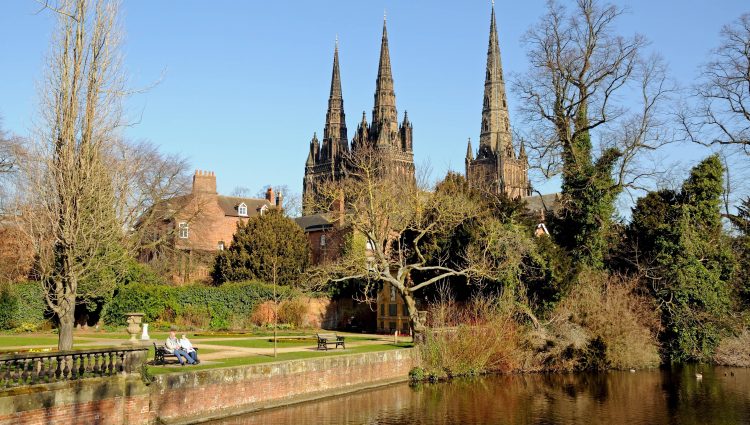 Landscape image of Lichfield Cathedral in Staffordshire. The photo is taken from the other side of the river. Two people are sat on a wooden park bench in front of the cathedral, and large trees frame the cathedral.