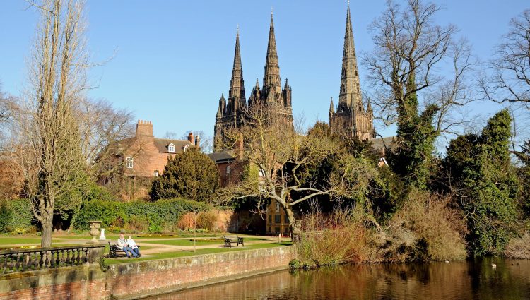 Landscape image of Lichfield Cathedral in Staffordshire. The photo is taken from the other side of the river. Two people are sat on a wooden park bench in front of the cathedral, and large trees frame the cathedral.