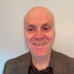 Head shot of Matt Harrison, Engagement Manager for East Midlands. Matt is standing against a white wall and smiling at the camera. He is wearing a black shirt and a blazer.