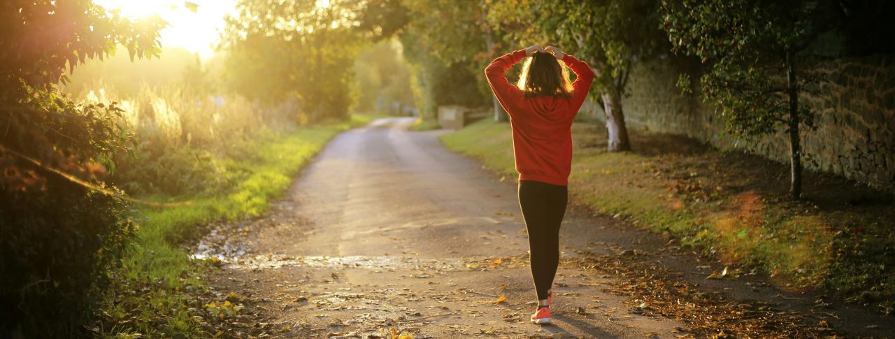 A female in gym gear, walking down a tree-lined, country lane, at sunset. She has her back to the camera, and her hands on her head.