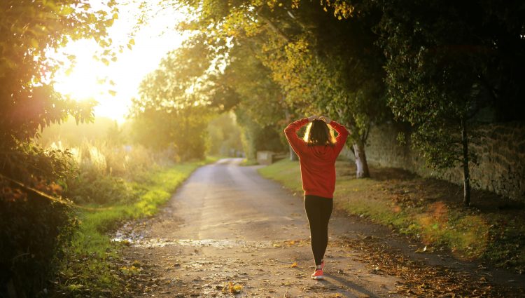 A female in gym gear, walking down a tree-lined, country lane, at sunset. She has her back to the camera, and her hands on her head.