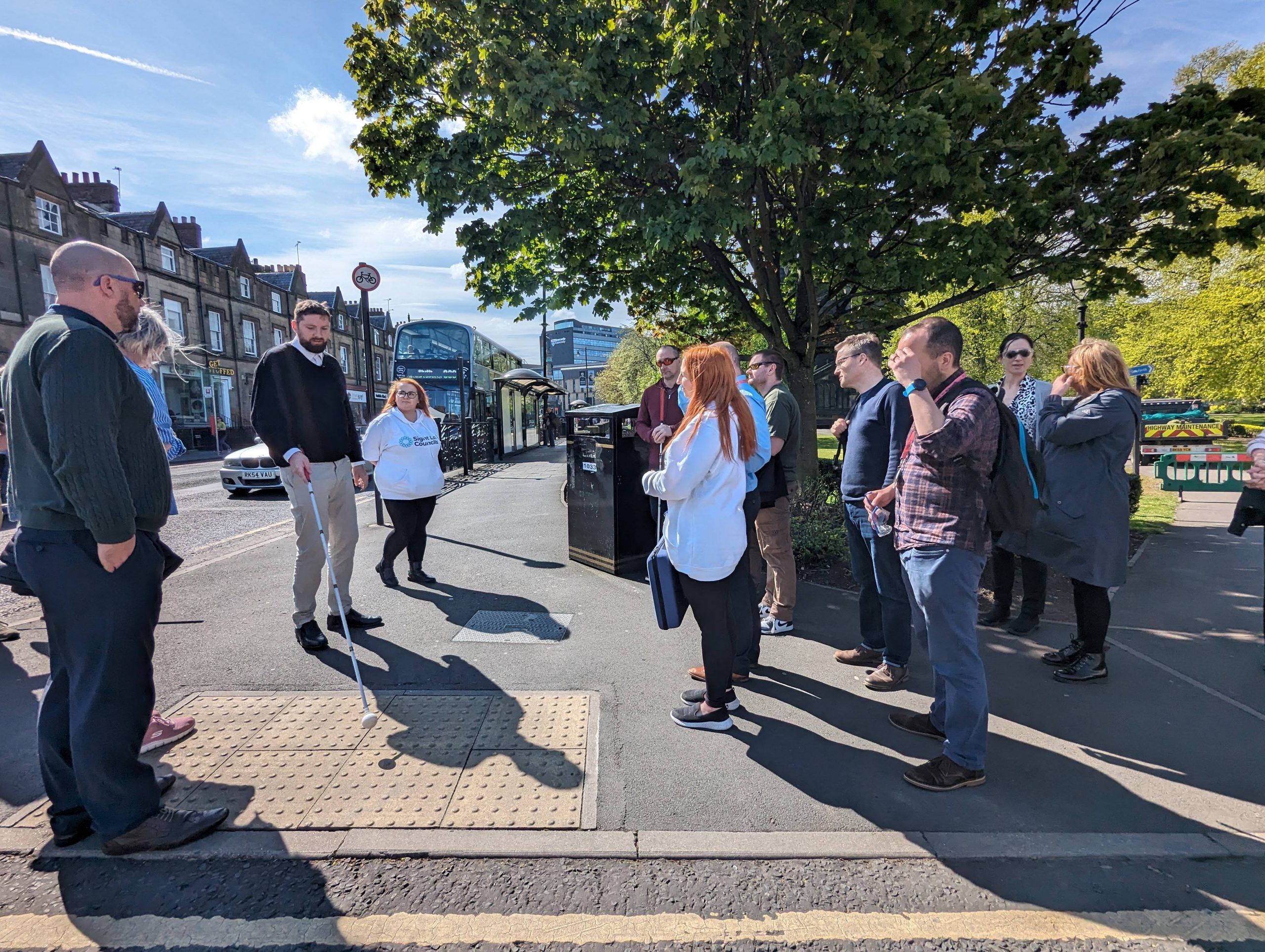 Jack, Engagement Manager for North East England, standing with officers from Newcastle City Council next to tactile markings on a pavement. He is holding his cane above the tactile marking as he talks to them.