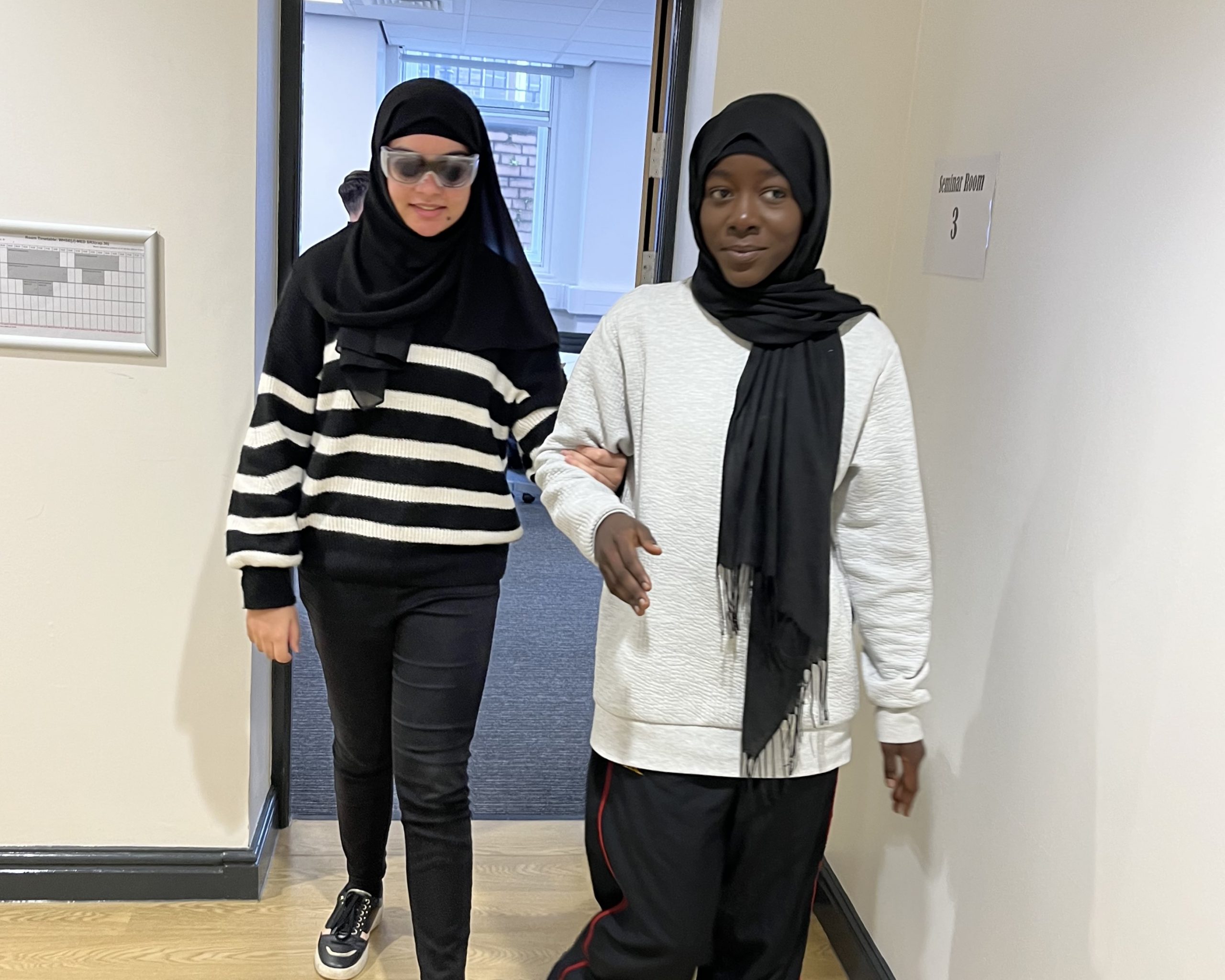 Two female medical students during the sight guide training. One is wearing sim specs, and the other is guiding their colleague through a doorway.