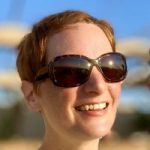 Headshot of Karin, Tyne and Wear SLC member. Helen has short, dark hair and is wearing a green t-shirt and sunglasses. The sun is shining on her face as she looks in the distance, she is smiling.