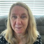Photo of Helen Stain, Derbyshire SLC member. Debbie has shoulder length, fair hair, and is wearing a black top with white flowers. She is sitting down in front of a window, smiling at the camera.