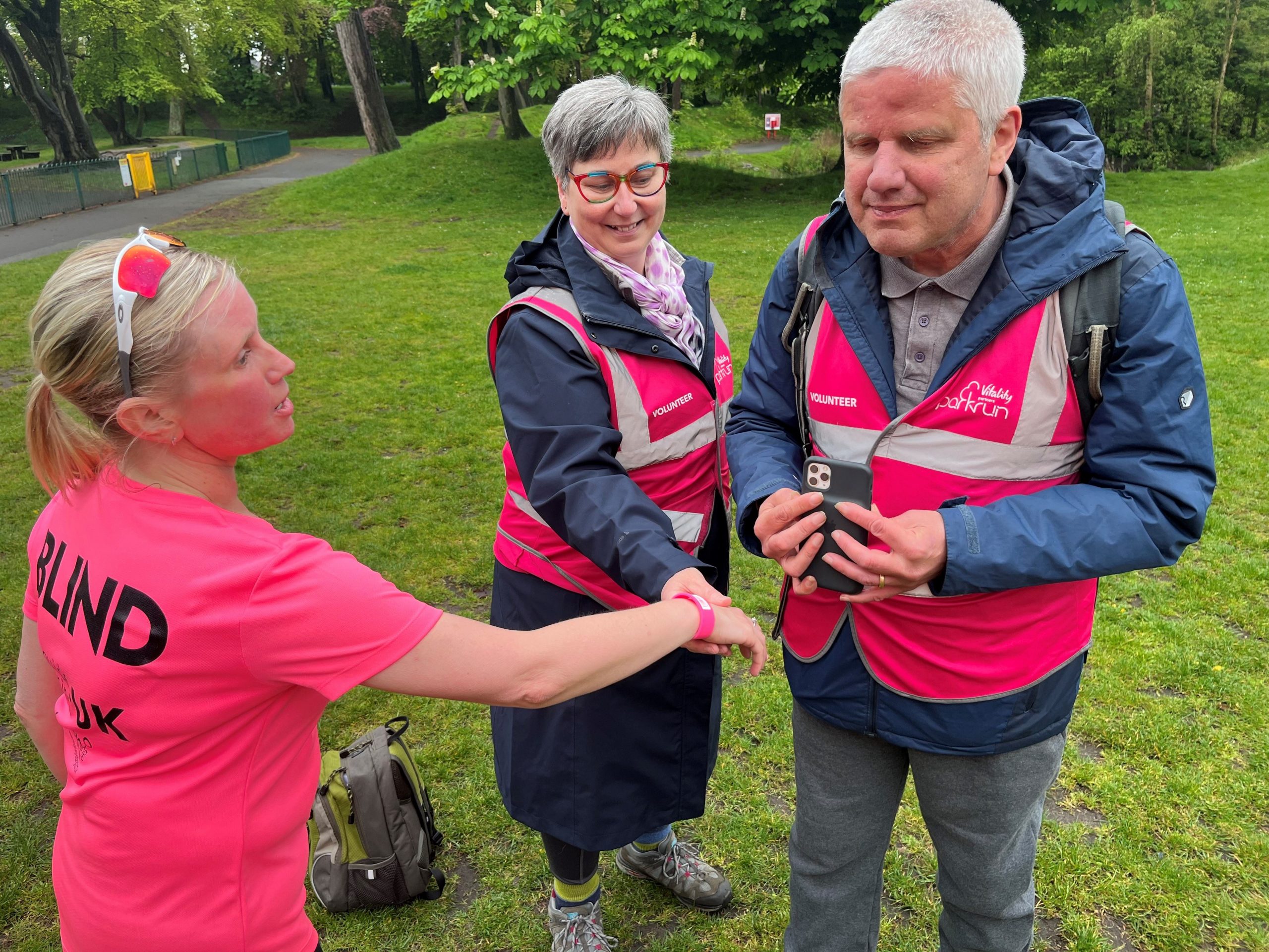 Kelly Barton, Engagement Manager for North West England at parkrun. She is having her barcode scanned by Merseyside SLC volunteer, Mike Wright Merseyside. Another parkrun volunteer is standing beside them.