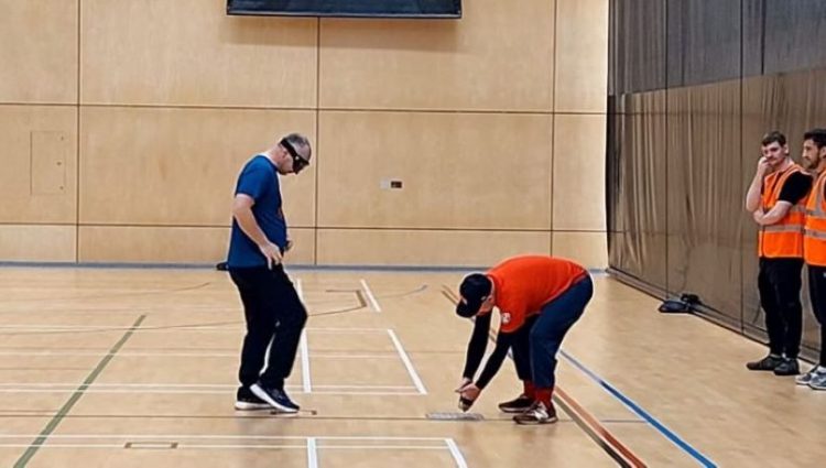There are four people in a sports hall. A man in a blue top and black trousers has back facing a net and is standing in front of another man in a red top who is knelt a few feat away from him and placing a ball on the ground. There are two people in orange tabards standing against a black wall behind them.
