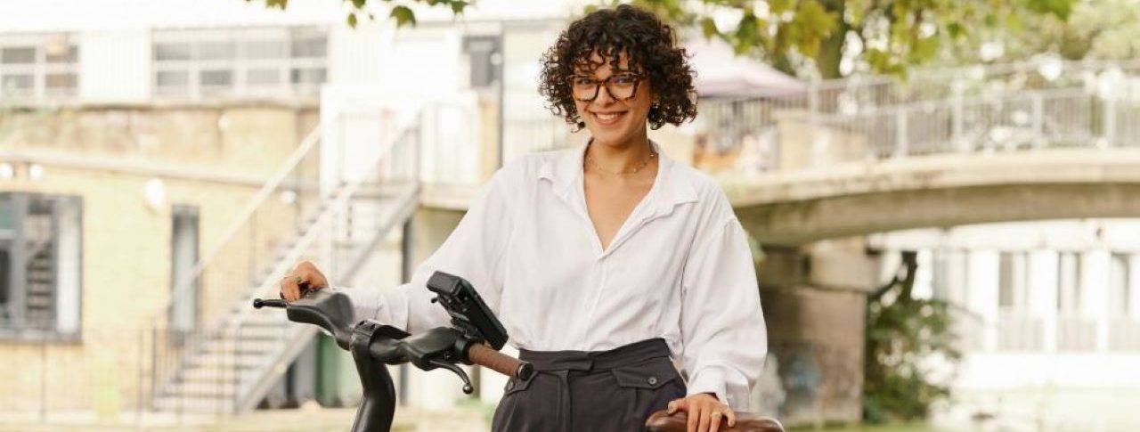 Woman dressed in smart attire posing with a dark green Forest bike on a leafy street. She is dressed in a white shirt and long black trousers and has short black hair.