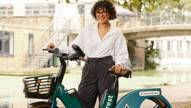 Woman dressed in smart attire posing with a dark green Forest bike on a leafy street. She is dressed in a white shirt and long black trousers and has short black hair.