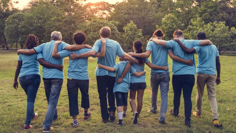 Group of volunteers with their arms around each other, facing away from the camera. They are outside and are all wearing light blue t-shirts.