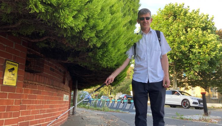 Dave Smith, Engagement Manager for South East England, is pictured next to a large hedge which is overhanging into the pavement. Dave is holding his long cane and looking at the camera.