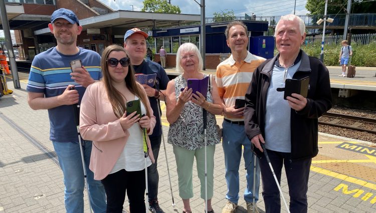 Members of Bedfordshire SLC are stood with Samantha Leftwich, Engagement Manager for East England. They are stood on a platform at Stevenage train station as part of the Aira trial. They are all holding their smartphones in one hand, their long cane in the other.