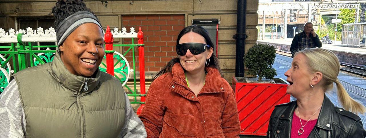 Three women at a train station. From left to right: a woman wearing a green gilet is guiding the woman standing beside her, who is wearing sim specs. Sight Loss Council Engagement Manager Kelly is standing on the right talking to them.