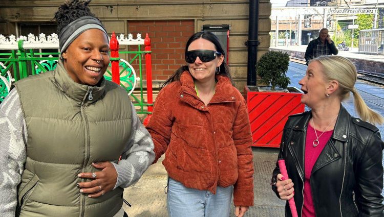 Three women at a train station. From left to right: a woman wearing a green gilet is guiding the woman standing beside her, who is wearing sim specs. Sight Loss Council Engagement Manager Kelly is standing on the right talking to them.