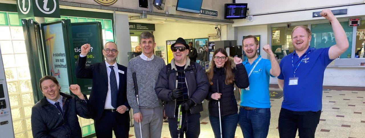Group photo of members testing the AIRA app at Chichester station. There are six people in the photo with some with their fists in the air, all smiling at the camera.