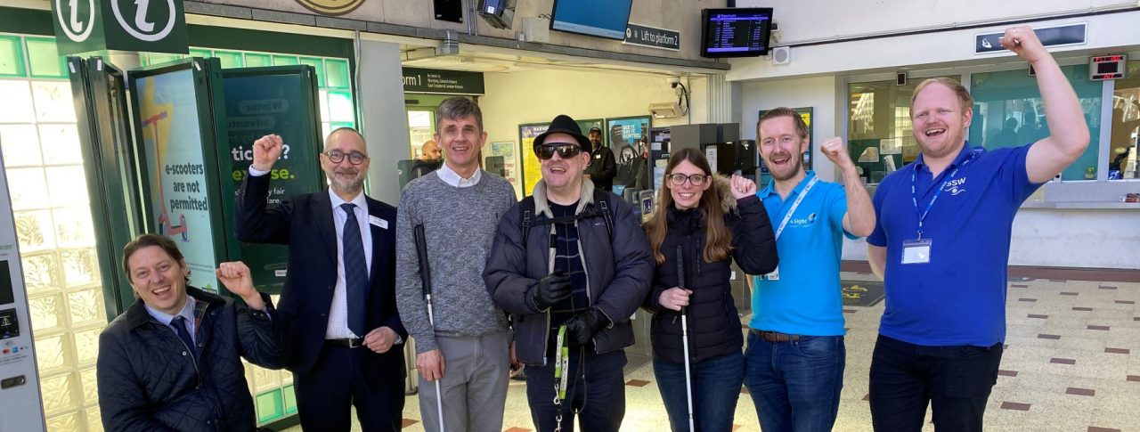 Group photo of members testing the AIRA app at Chichester station. There are six people in the photo with some with their fists in the air, all smiling at the camera.