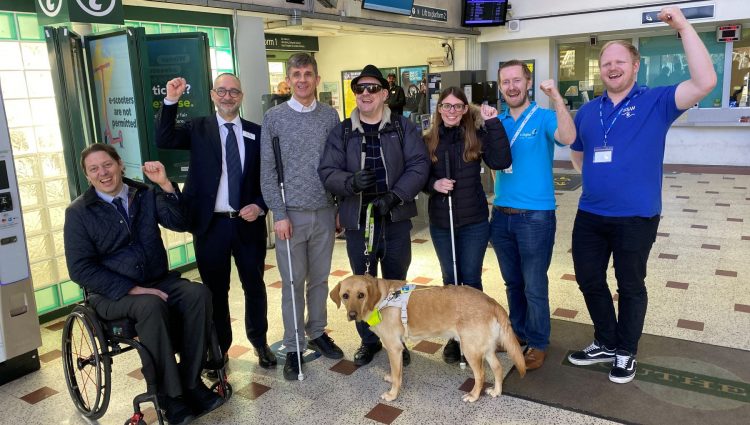 Group photo of members testing the AIRA app at Chichester station. There are six people in the photo with some with their fists in the air, all smiling at the camera.