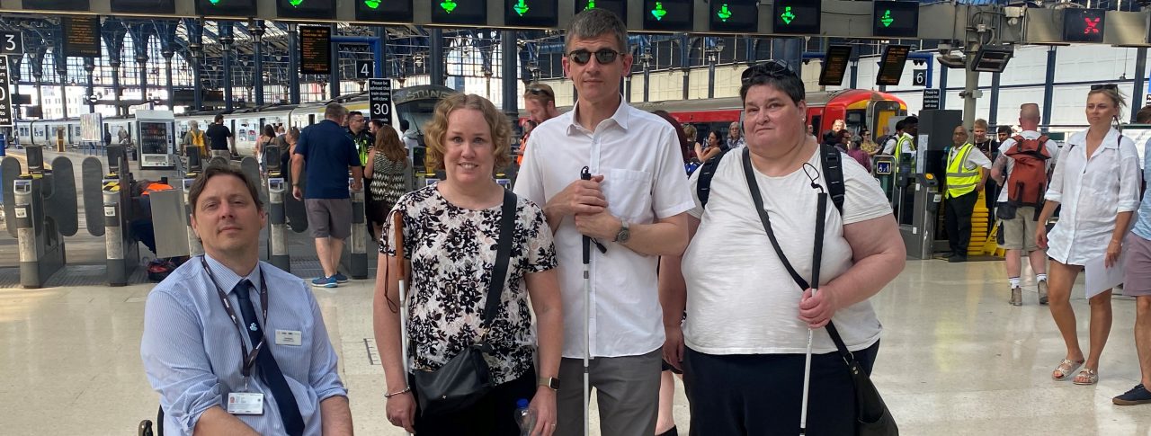 From left to right: Carl Martin, Accessibility Lead for Govia Thameslink Railways; Linn Davies, East Sussex SLC member; Dave Smith, Engagement Manager for the South East, and Iris Keppler, East Sussex SLC member. They are in the concourse of Brighton train station, looking at the camera, smiling.
