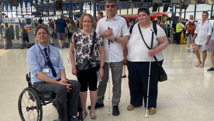 From left to right: Carl Martin, Accessibility Lead for Govia Thameslink Railways; Linn Davies, East Sussex SLC member; Dave Smith, Engagement Manager for the South East, and Iris Keppler, East Sussex SLC member. They are in the concourse of Brighton train station, looking at the camera, smiling.