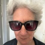 Steph Syson headshot. Steph has short curly grey hair and is wearing dark glasses and a black t-shirt.