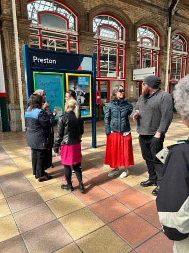 Seven people standing outside a sign for Preston railway station. All appear to be engaged in conversation, with one female attendee wearing sim specs.