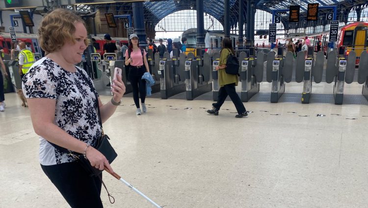 SLC volunteer Linn walking through a train station using the Aira app. She has her smartphone in one hand and her white navigation cane in the other.