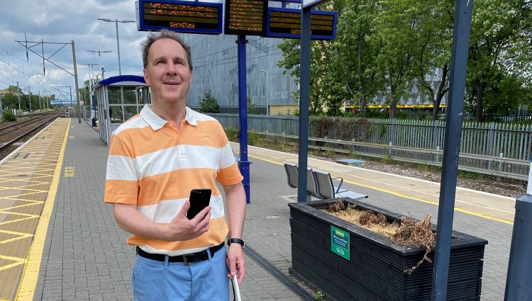 Paul Day, Bedfordshire SLC member, is standing on a platform at Stevenage train station. he is holding his long cane in one hand, and his phone up in the other.