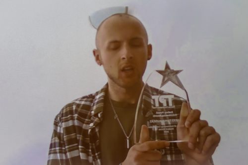 Aleks is standing facing the camera, holding up his award with both hands. He is wearing a striped shirt over the top of a khaki t-shirt and a silver necklace.