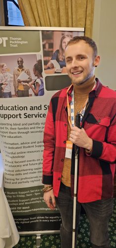 Student-Voice volunteer Aleks posing next to a TPT banner, which is positioned to the left. Aleks has light brown hair, wearing a red rain jacket and holds a white cane in his right hand. 