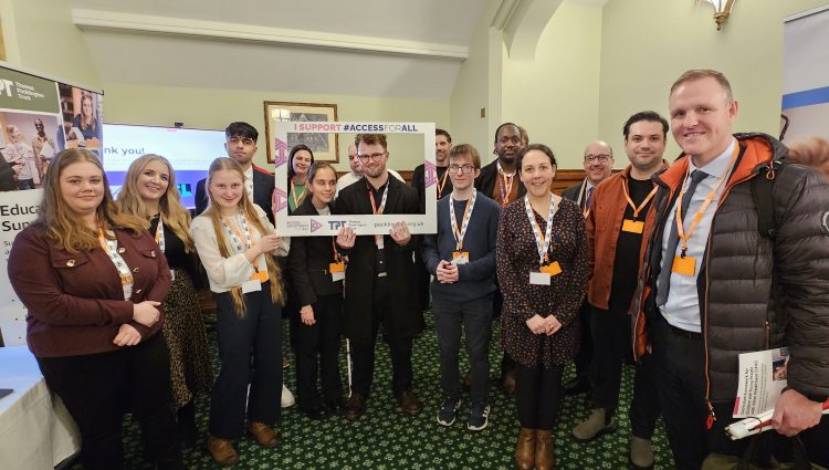 A group of around 15 people stand closely together indoors, smiling at the camera. In the centre, two people are holding a large cardboard photo frame that reads 'I support #ACCESSFORALL'. The Thomas Pocklington Trust logo and website are clearly visible at the bottom of the frame.