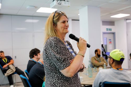 TPT Director of Services at Thomas Pocklington Trust speaks into the microphone at the SLC conference. Delegates are sat in the background around round tables.