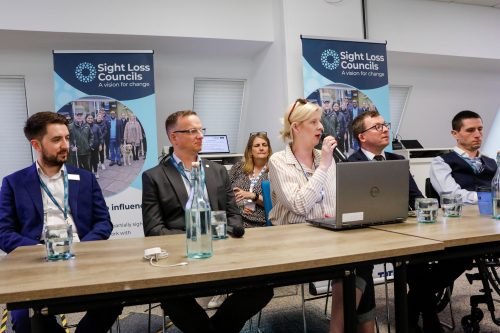 Male representatives from the Rail Delivery Group, National Rail, Confederation for Passenger Transport and Driver and Vehicle Standards Agency sat at a top table infront of two SLC banners for a question and answer panel. Louise Connop is sat in the centre speaking into a microphone.