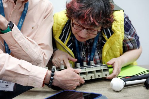 A SLC volunteer is exploring a tactile tool used to hold money in buses. Their white navigation cane is next to the on the table.