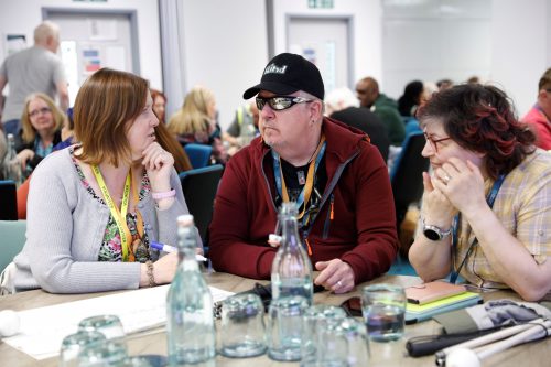 Two SLC volunteers and a sighted guide engage in discussions at a round table. The guide is making notes on flip chart. A navigation cane is laid on the table. Other volunteers are sat around tables in the background.
