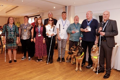 Group photo of ten people and two guide dogs. They are all smartly dressed, four of them are holding canes, and they are all wearing a medal. The two guide dogs are to the right, one is an Alsatian and one is a Labrador.