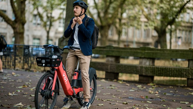 White man fastens a black helmet while sitting on an orange Voi e-bike, preparing to cycle off. He is smartly dressed in beige chinos and a navy jacket.