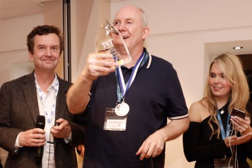 CEO Charles, Paul, and Claire Sisk. They are all dressed smartly and look happy. Charles is on the left and is holding a microphone. Paul is in the middle and is holding his award up to the camera, and is also wearing a medal. Claire is on the right, looking at Paul, and is holding her mobile phone.