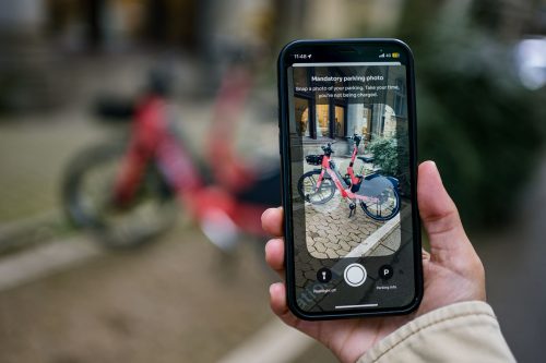 Focused photograph foregrounding a hand holding a mobile phone. The phone shows Voi's mandatory parking photo screen, with an orange Voi e-bike parked in a designated parking space. 